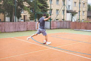 Obraz premium Young man takes a powerful shot on goal while playing football on an outdoor court. Wearing casual attire, he shows intensity and focus in a dynamic moment with an urban backdrop behind him