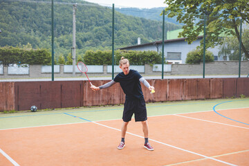 Fototapeta premium Badminton player stands ready to serve on an outdoor court, holding a racket and shuttlecock. Dressed in casual sportswear, he focuses on the game amid a scenic, natural setting
