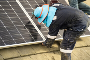 Builder mounting photovoltaic solar modules on roof of house. Back view of man engineer in helmet installing solar panel system outdoors. Concept of alternative and renewable energy.