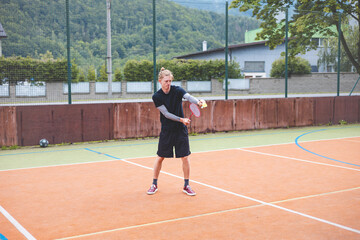 Badminton player stands ready to serve on an outdoor court, holding a racket and shuttlecock. Dressed in casual sportswear, he focuses on the game amid a scenic, natural setting