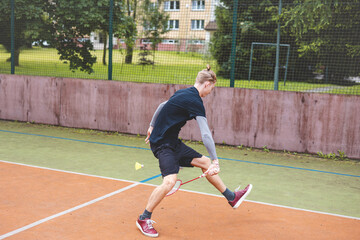 Badminton player makes an agile, low return shot on an outdoor court, showcasing flexibility and quick reflexes. Dressed in casual sportswear, he focuses on hitting the shuttlecock with precision