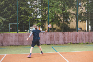 Badminton player strikes the shuttlecock during a game on an outdoor court, showcasing precise technique and athletic movement. The player is focused, surrounded by trees and an urban backdrop