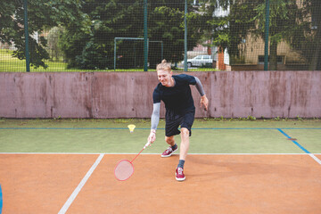 Badminton player strikes the shuttlecock during a game on an outdoor court, showcasing precise technique and athletic movement. The player is focused, surrounded by trees and an urban backdrop