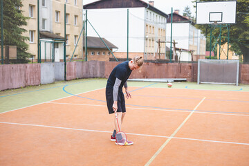 Badminton player picks up a shuttlecock during a game on an outdoor court. The casual moment takes place in an urban setting, with surrounding buildings and a basketball hoop in the background