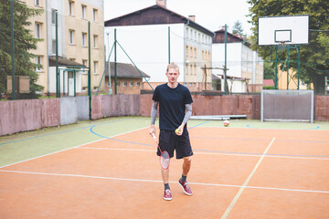 Obraz premium Badminton player picks up a shuttlecock during a game on an outdoor court. The casual moment takes place in an urban setting, with surrounding buildings and a basketball hoop in the background