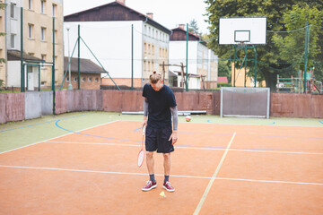 Fototapeta premium Badminton player picks up a shuttlecock during a game on an outdoor court. The casual moment takes place in an urban setting, with surrounding buildings and a basketball hoop in the background