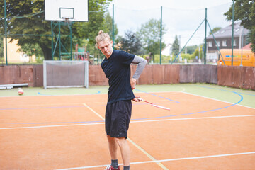 Badminton player strikes the shuttlecock during a game on an outdoor court, showcasing precise technique and athletic movement. The player is focused, surrounded by trees and an urban backdrop