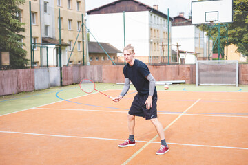 Badminton player focuses on the shuttlecock mid-air during a match on an outdoor court. With his racket raised, he prepares for the next shot, surrounded by an urban and scenic background