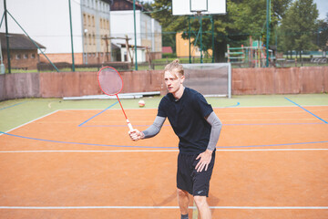 Obraz premium Badminton player strikes the shuttlecock during a game on an outdoor court, showcasing precise technique and athletic movement. The player is focused, surrounded by trees and an urban backdrop
