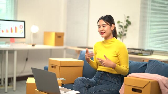 Asian woman entrepreneur is sitting on a couch and speaking on a video call on her laptop, with cardboard boxes next to her