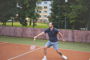 Badminton player focuses on the shuttlecock mid-air during a match on an outdoor court. With his racket raised, he prepares for the next shot, surrounded by an urban and scenic background