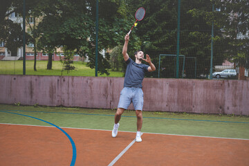 Badminton player strikes the shuttlecock during a game on an outdoor court, showcasing precise technique and athletic movement. The player is focused, surrounded by trees and an urban backdrop
