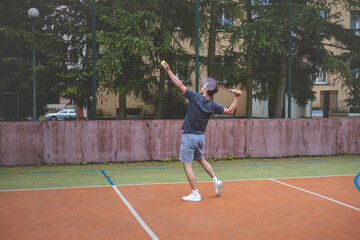 Fototapeta premium Badminton player strikes the shuttlecock during a game on an outdoor court, showcasing precise technique and athletic movement. The player is focused, surrounded by trees and an urban backdrop