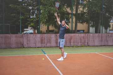 Badminton player strikes the shuttlecock during a game on an outdoor court, showcasing precise technique and athletic movement. The player is focused, surrounded by trees and an urban backdrop