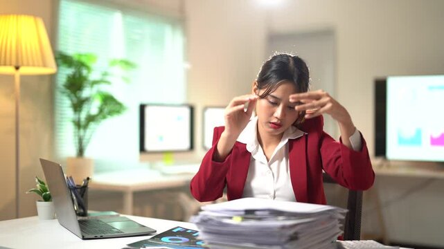 Young businesswoman in a red blazer is working late, struggling to stay awake as she battles a mountain of paperwork at her desk