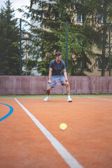 Badminton player celebrates a moment on an outdoor court, holding his racket in an energetic stance. His expression shows excitement and triumph, set against a backdrop of trees and buildings