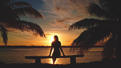 Woman sitting on a bench between palm trees, gazing at a stunning sunset on a tropical island in french polynesia's maupiti
