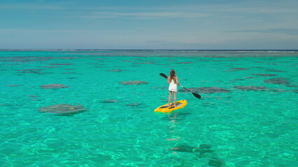 Woman paddleboarding in tropical waters, enjoying vacation in paradise. Crystal-clear turquoise sea, sunny sky, and coral reefs visible below. Perfect getaway for relaxation and fun