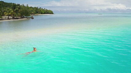 Aerial view of a woman swimming in the turquoise water of a tropical lagoon. The lagoon is surrounded by a white sand beach and palm trees