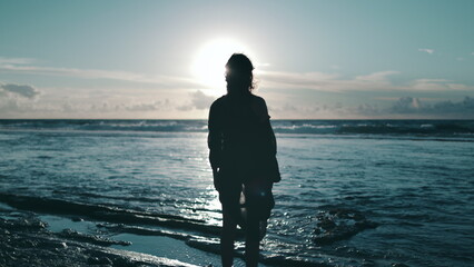 A person stands alone on a sandy beach next to the roaring ocean waves, staring out towards the...