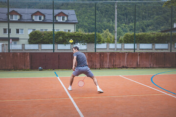 Badminton player returns a shot on an outdoor court, extending his body for a powerful hit. The scene captures athletic focus, with a background of buildings and greenery surrounding the court