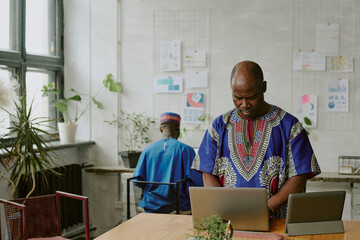 Mature African American programmer using tablet and laptop while hi African African colleague sitting at work table in background