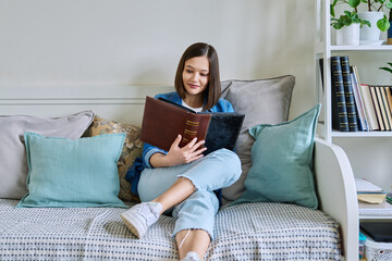 Young happy woman looking at photo album sitting on couch at home