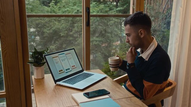 Medium shot of Caucasian man enjoying his morning coffee while deciding on subscription plan on laptop, sitting by window at home desk