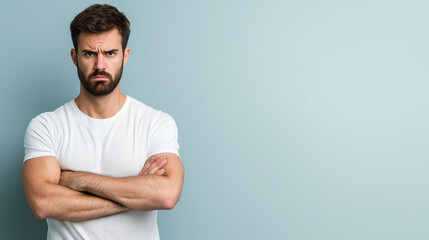 Fototapeta premium A man with furrowed brows, arms crossed tightly across his chest, conveying a sense of suspicion, standing against a pastel gray background.