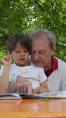 Grandfather and grandson reading a book together outdoors, under the shade of a tree, enjoying quality time and fostering a strong family bond and intergenerational learning