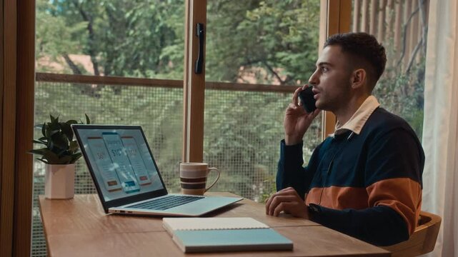 Side view of Caucasian businessman choosing subscription plan on laptop while discussing on smartphone at home desk