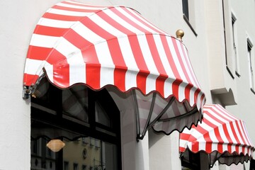 Rot und weiß gestreifte, runde Markisen vor einem Geschäft. Die Gebäude auf der gegenüberliegenden Straßenseite spiegeln sich im Schaufenster.