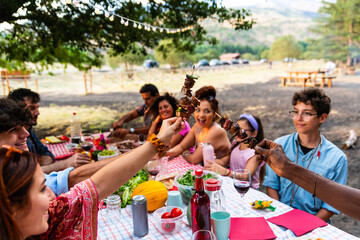 toasting with meat skewers - diverse group of friends celebrating grilled meat skewers at outdoor barbecue – multiracial young people having fun at park barbecue grilling and eat meat skewer