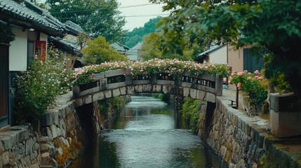 Fototapeta premium A simple stone pedestrian bridge, crossing a narrow river in a small village, with flower-lined banks on both sides