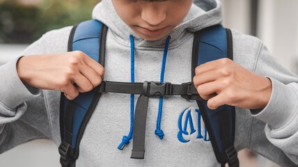 Close-up view of hands fastening a backpack strap. The image showcases a person wearing a grey hoodie, adjusting the chest strap of a dark-colored backpack.