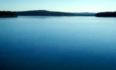 Fototapeta premium A serene lake, surrounded by a forested landscape, stretches out towards the horizon under a clear blue sky