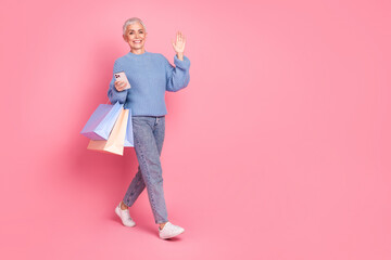 Full length photo of pretty retired female walk wave hand shopping device wear trendy blue knitwear outfit isolated on pink background
