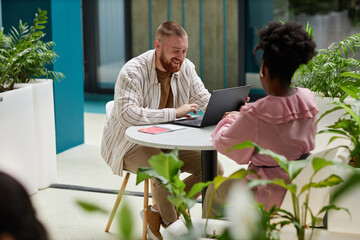 Full length shot of smiling adult man using laptop sitting at cafe table in office center while having work related conversation with female colleague, copy space