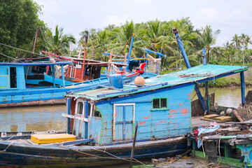 Colorful fishing boats docked along the concrete pier at Pelabuhan Kuala Singkawang, Kalimantan Barat, with village buildings and lush trees in the background, capturing coastal life.