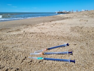 Syringes in the sand with a city skyline in the distance