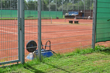 View of Outdoor Clay Tennis Courts with Upturned Wheelbarrow seen through Steel Fence