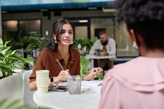 Medium shot of businesswoman talking to female colleague at business meeting discussing work strategy while sitting at cafe table in contemporary office center, copy space