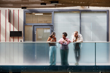 Wide angle shot of diverse group of three colleagues cheerfully chatting during break while standing on balcony in sunlight casting through glass ceiling in modern business center, copy space © Seventyfour