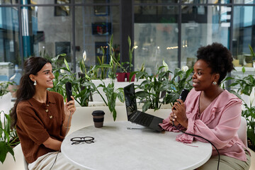 Two smiling women engaged in lively discussion about feminism co hosting podcast episode. Two successful businesswomen recording interview in cafe, copy space