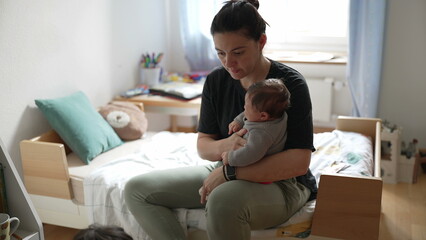 Mother sitting on a child’s bed in a tidy and bright children’s bedroom, holding her baby while her young son plays on the floor nearby, capturing a candid and authentic family lifestyle