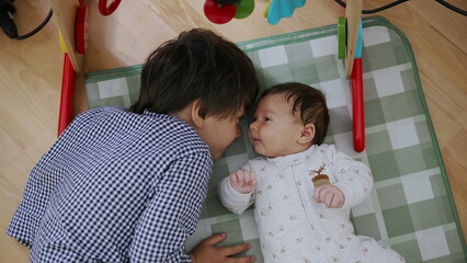 Older brother lying next to his baby sibling on a play mat, sharing a loving moment and gazing into...