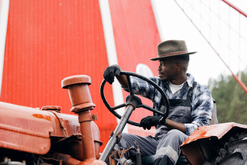 Happy African American young man tractor driver preparing to harvest wheat, hay against background of mill. Concept farmer agriculture industry