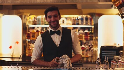 Profile good looking smiling smart bartender wiping glass at luxurious beverage counter bar scene looking at camera to pose neat professional barman costume greeting warm at bar nightclub. Vinosity.