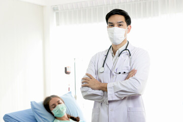 Portrait of young male doctor with stethoscope wears face mask, stands with arms crossed in white treatment room with the patient lying down on bed as a blurred background. People in the medical field