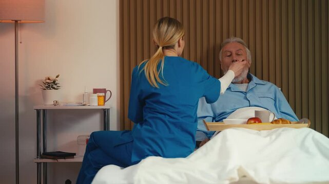Caring nurse spoon-feeding elderly male patient, home care, assisted living